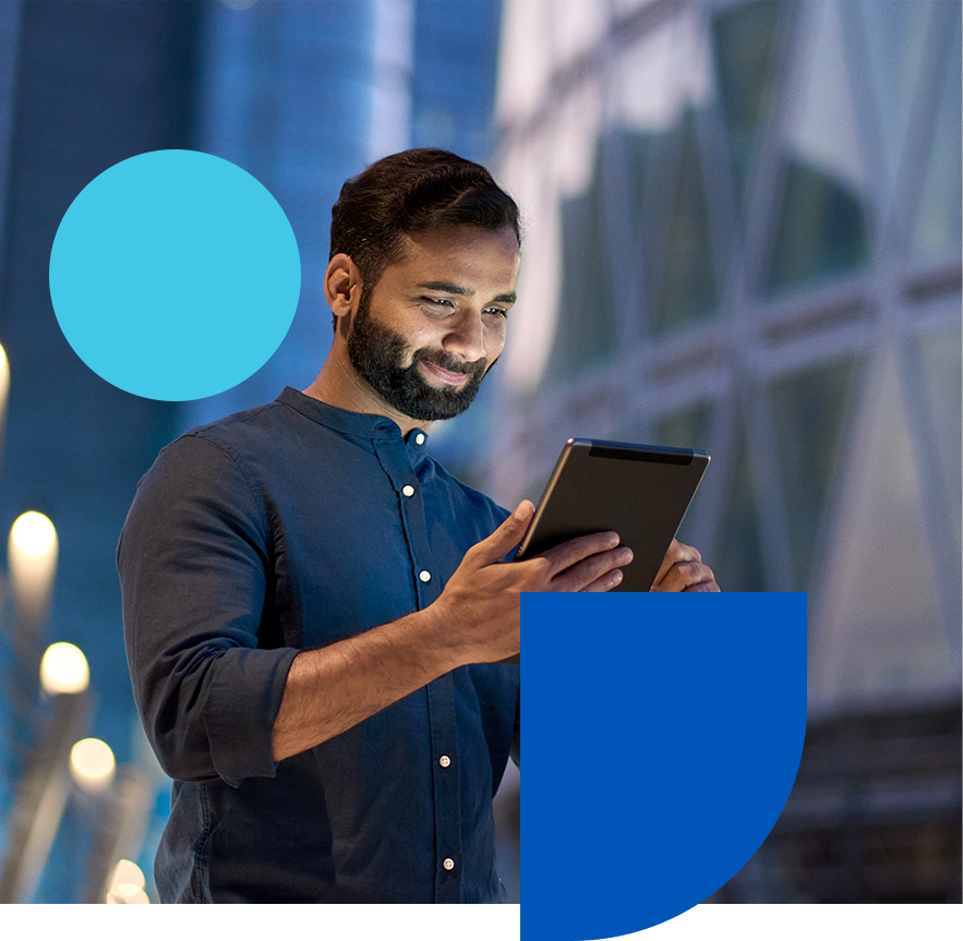 A man in a dark shirt is smiling while using a tablet. He stands in a modern urban setting with blurred lights in the background. Geometric shapes in blue are overlaid on the image.